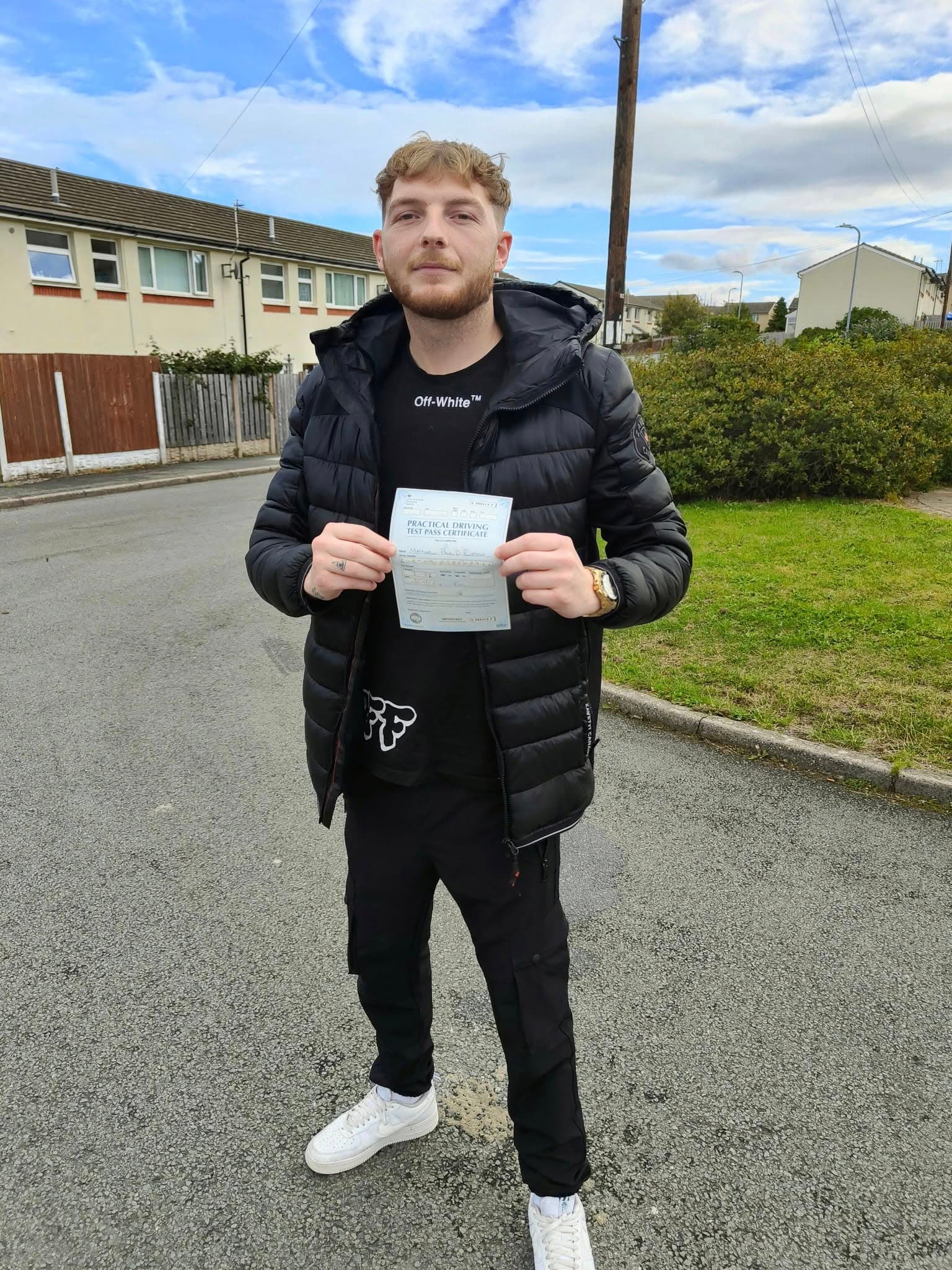 Young man in black puffer jacket holding a practical driving test pass certificate outdoors.