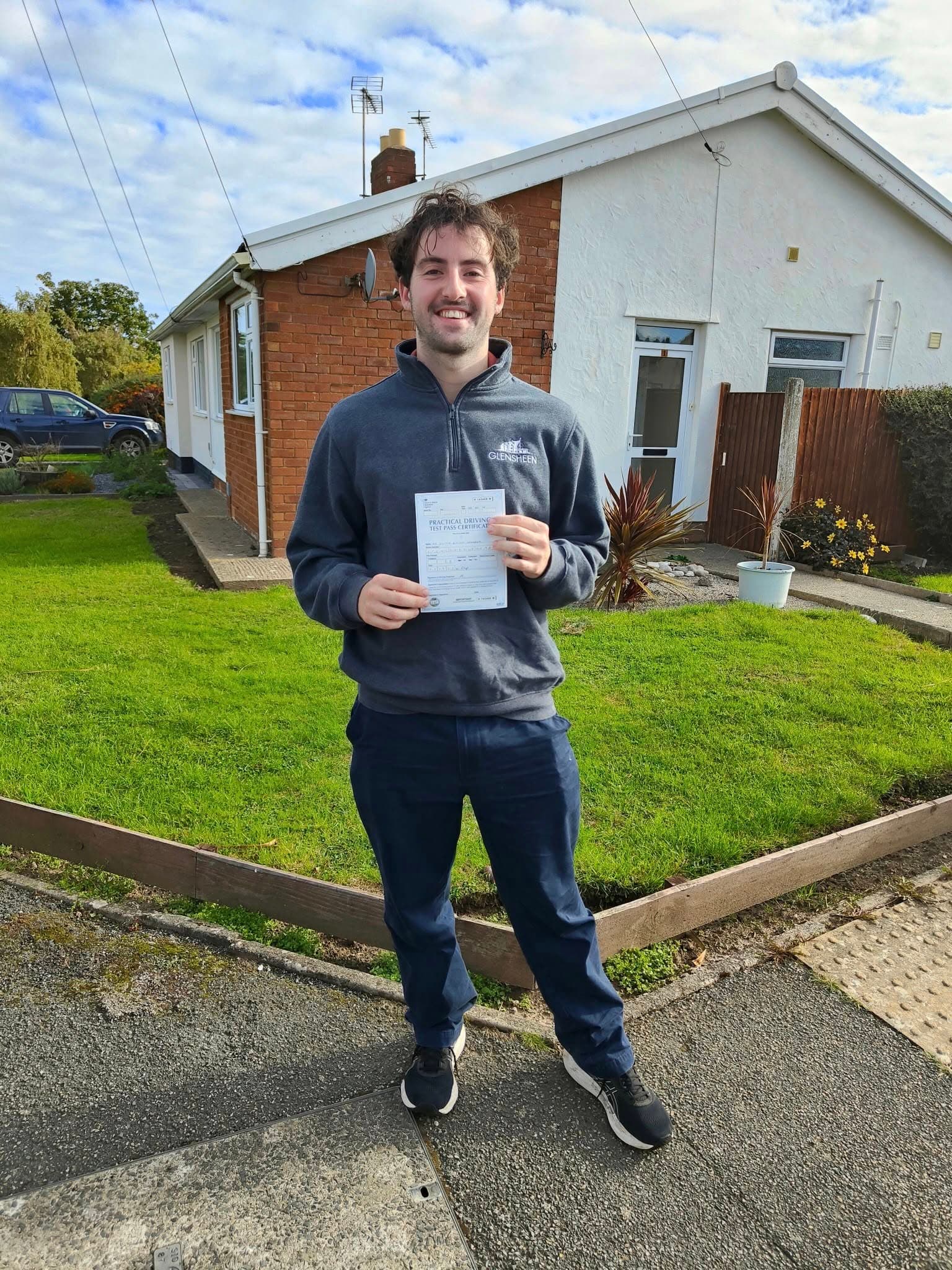 Smiling man stands outside a house holding his practical driving test pass certificate.